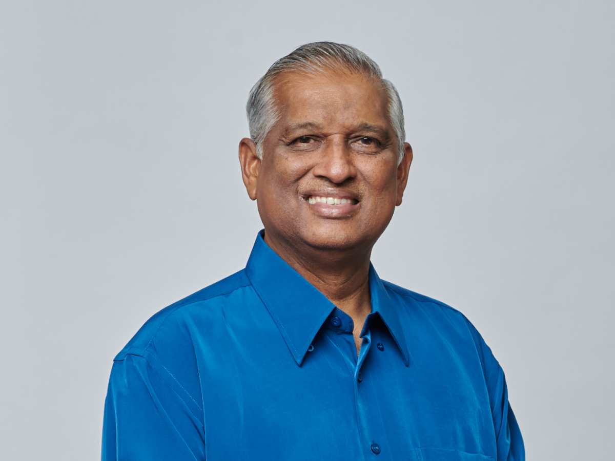 Mr Naseer Bin Ghani in a bright blue shirt smiling with hands clasped in front against a plain grey background